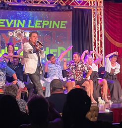 Live stage hypnosis show with a male performer in a white jacket holding a microphone as seated volunteers mimic flexing muscles under colorful stage lights, audience silhouettes in foreground.