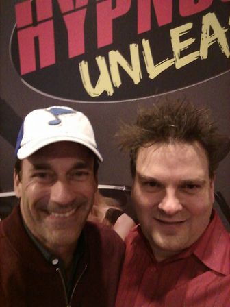 Selfie of two smiling men at an indoor event in front of a colorful promotional backdrop reading UNLEA... — one wearing a white baseball cap, the other in a red shirt