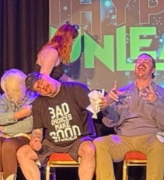 Three people seated on an indoor comedy-night stage — two men laughing and one wearing a T-shirt that reads "Bad choices make good stories", colorful backdrop and stage lighting.