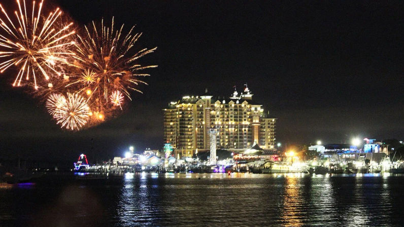 Nighttime fireworks bursting over a brightly lit waterfront hotel and pier, colorful reflections shimmering on the harbor water.