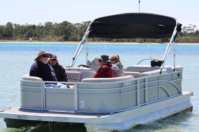 Four adults relaxing on a pontoon boat with a black canopy in clear turquoise water near a tree-lined shoreline on a sunny day