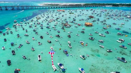 Aerial view of a crowded tropical sandbar with dozens of boats, swimmers and floating bars in clear turquoise water near a coastal bridge