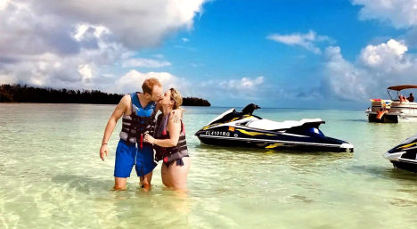 Couple in life jackets kissing waist-deep in clear turquoise shallow water beside a parked jet ski and a pontoon boat under a bright blue sky with fluffy clouds — tropical beach water-sports scene