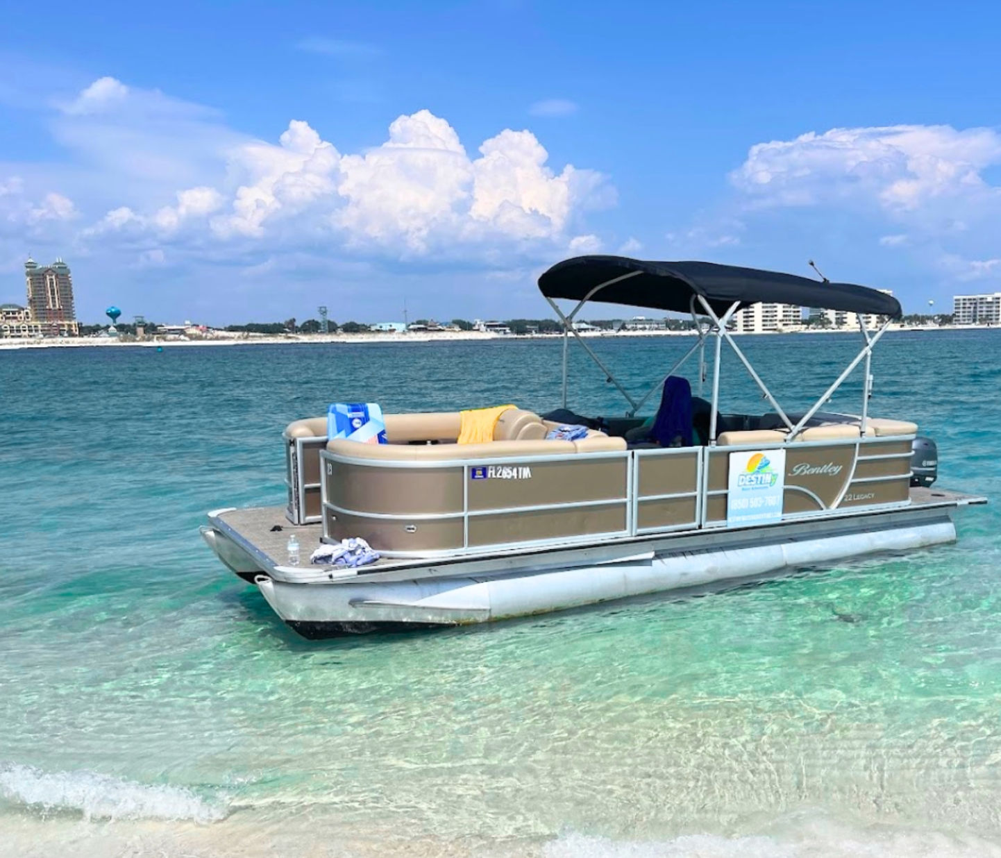 Pontoon boat with black canopy anchored in clear turquoise water off a sandy beach, distant beachfront buildings on the horizon and a bright blue sky with puffy white clouds — a sunny coastal day.