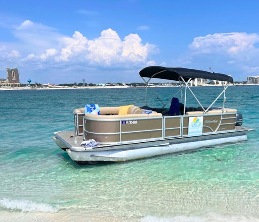 Pontoon boat with black canopy anchored in clear turquoise water off a sandy beach, distant beachfront buildings on the horizon and a bright blue sky with puffy white clouds — a sunny coastal day.