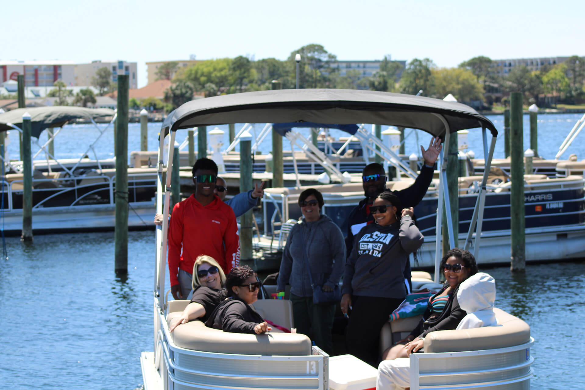 Pontoon boat at a sunny marina with a group of friends lounging, smiling and waving near docks and moored boats on blue water.