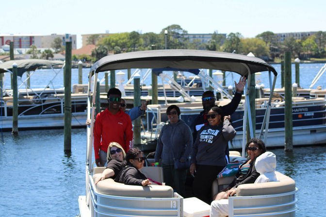 Pontoon boat at a sunny marina with a group of friends lounging, smiling and waving near docks and moored boats on blue water.