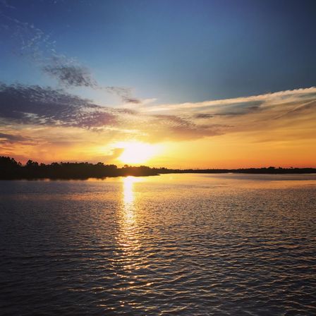 Vibrant sunset over a calm river with golden sunlight reflecting on rippling water, silhouetted tree-lined horizon, and colorful orange and blue sky.