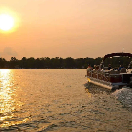 Pontoon boat cruising across a golden lake at sunset, sun low in the sky casting warm reflections on rippled water with a tree-lined shoreline in the distance.