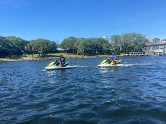 Two riders on bright yellow-green jet skis cruising a sunny, tree-lined residential waterway with docks and clear blue sky