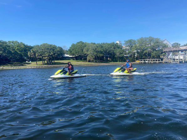 Two riders on bright yellow-green jet skis cruising a sunny, tree-lined residential waterway with docks and clear blue sky