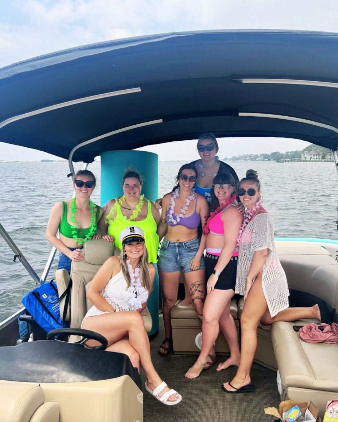 Seven friends in colorful swimsuits and leis posing on a covered pontoon boat near a bay shoreline for a sunny summer boat party, one wearing a captain’s hat