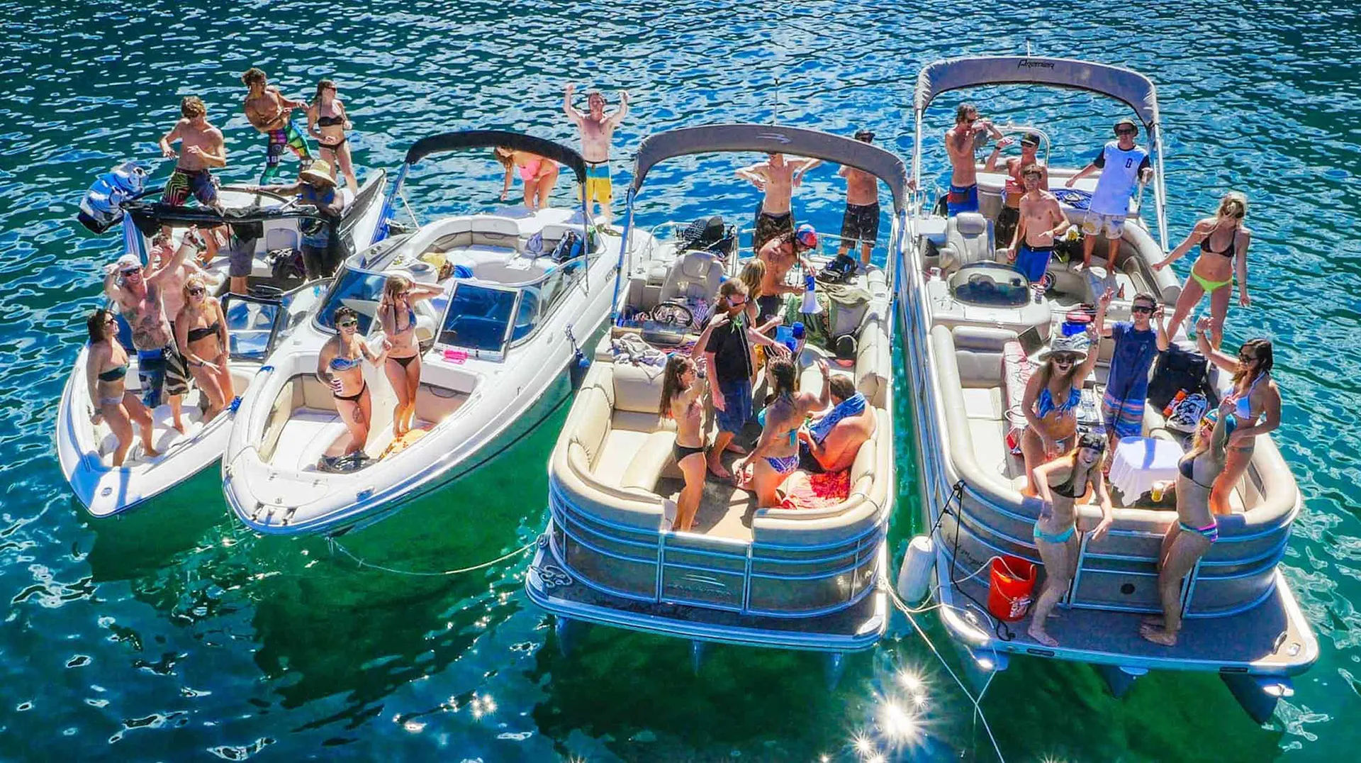 Aerial view of four boats tied together on a sunny lake with groups in swimsuits partying on pontoons and speedboats, sun sparkling on green-blue water.