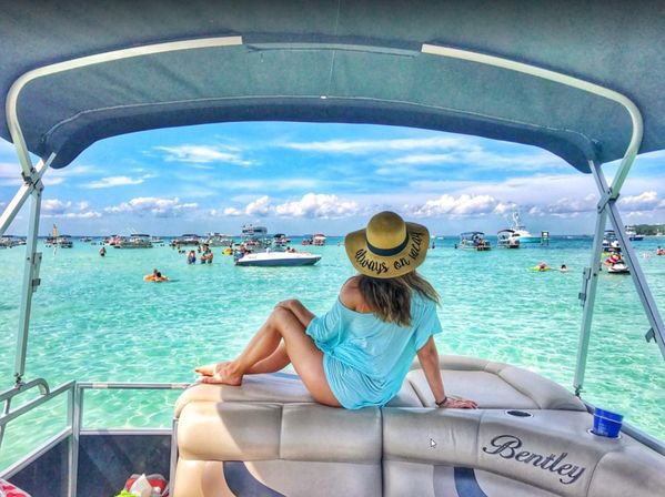 Woman in a straw sun hat lounging on the stern of a pontoon boat, gazing at turquoise shallow water dotted with anchored boats and swimmers under a bright blue sky.