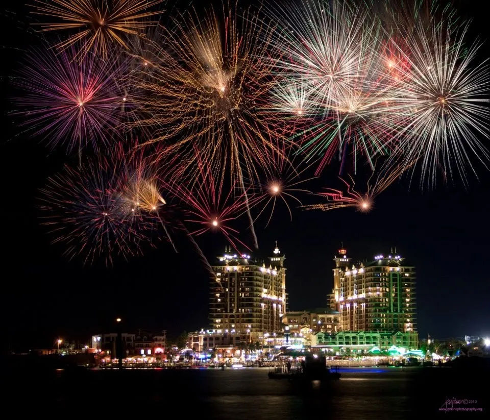 Vibrant multicolored fireworks exploding over a lit waterfront city skyline and harbor at night, high-rise buildings glowing and reflecting on the water
