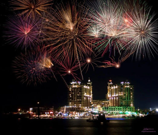 Vibrant multicolored fireworks exploding over a lit waterfront city skyline and harbor at night, high-rise buildings glowing and reflecting on the water
