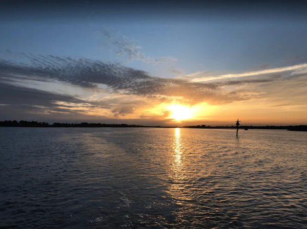Golden sunset over a calm coastal bay, sun reflecting on rippled water with a silhouetted navigation marker and distant tree-lined shoreline under dramatic clouds.