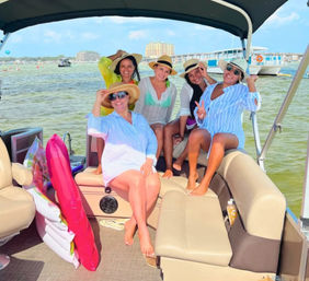 Five women in sun hats and cover-ups laughing on a pontoon boat with inflatables, coastal beach and skyline in the background on a sunny summer day