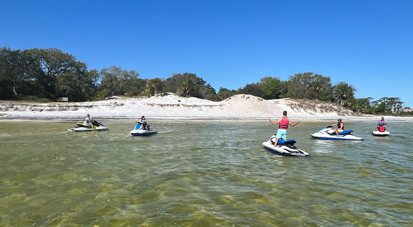 Five people on jet skis in shallow green coastal water with white sand dunes and trees under a clear blue sky