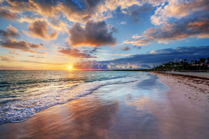 Tropical beach at sunset with golden sun, dramatic clouds, palm-lined shoreline and gentle ocean waves reflecting on wet sand.