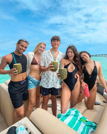 Five friends on a tropical boat in turquoise water, smiling in swimwear and holding hollowed pineapple drinks under a bright blue sky