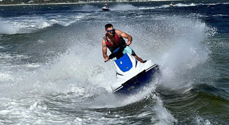 Man riding a blue-and-white jet ski carving through choppy coastal waters, spraying a dramatic wake with other personal watercraft visible near the shoreline.