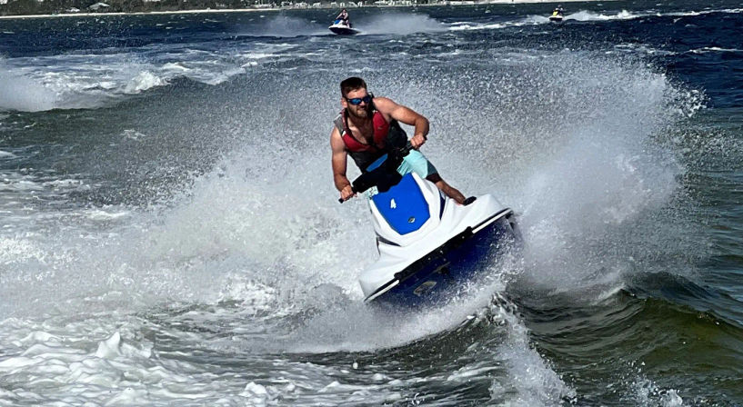 Man riding a blue-and-white jet ski carving through choppy coastal waters, spraying a dramatic wake with other personal watercraft visible near the shoreline.
