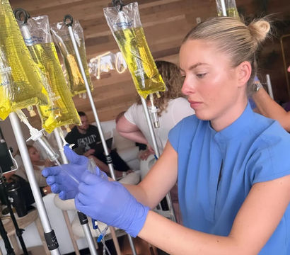 Person wearing blue gloves adjusts an IV pole with multiple hanging yellow infusion bags (vitamin drips) in a modern wellness clinic lounge.