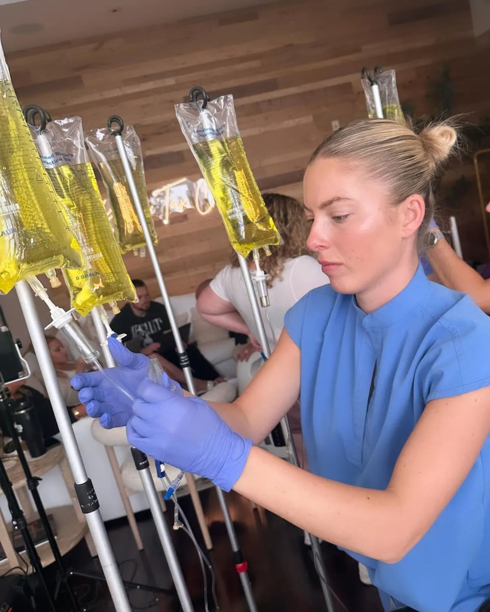 Person wearing blue gloves adjusts an IV pole with multiple hanging yellow infusion bags (vitamin drips) in a modern wellness clinic lounge.