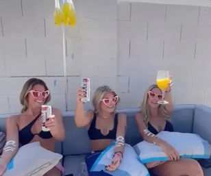 Three people in black bikinis and pink heart-shaped sunglasses cheering with cans and an orange cocktail while lounging on a sunny poolside patio couch with blue pillows and yellow balloons overhead