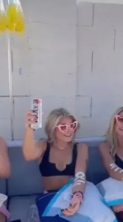 Three people in black bikinis and pink heart-shaped sunglasses cheering with cans and an orange cocktail while lounging on a sunny poolside patio couch with blue pillows and yellow balloons overhead