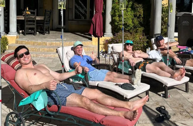 Four men lounging shirtless on poolside chaise lounges with IV drip poles, thumbs-up and sunglasses on a sunny backyard patio