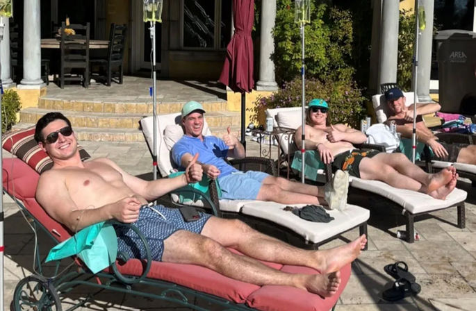 Four men lounging shirtless on poolside chaise lounges with IV drip poles, thumbs-up and sunglasses on a sunny backyard patio