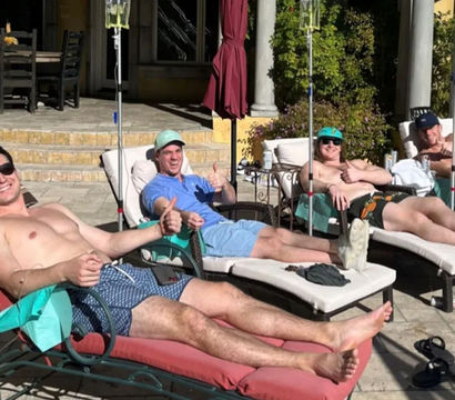 Four men lounging shirtless on poolside chaise lounges with IV drip poles, thumbs-up and sunglasses on a sunny backyard patio
