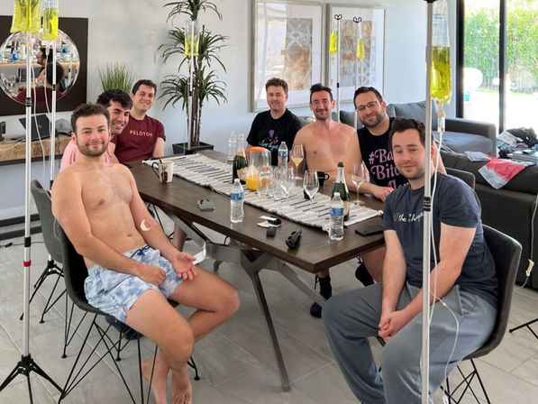 Seven men gathered around a dining table in a bright modern living room, some shirtless and hooked up to IV hydration drips from poles, with water bottles, glasses and bottles on the table — casual at-home wellness gathering.