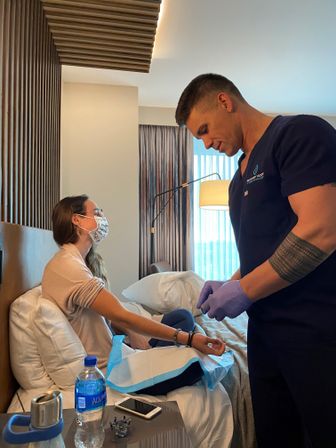 Healthcare worker in scrubs and purple gloves preparing a bedside medical treatment for a masked patient sitting on a hotel bed, with water bottle and phone on the bedside table.