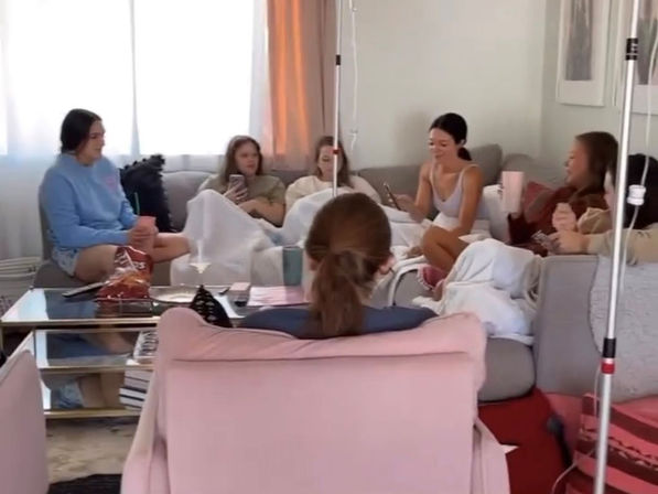 Cozy living room scene with a group of friends lounging on sofas under blankets, scrolling phones and snacking at a coffee table, with IV poles visible at the edges.