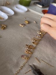 DIY jewelry close-up of a hand attaching gold charms and small pearl beads to a gold chain bracelet on a fabric tabletop, with pliers, headpins and small trays of charms nearby.