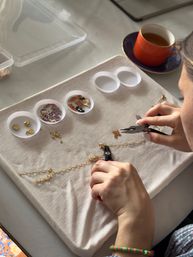 DIY jewelry-making scene: hands using pliers to assemble a gold chain charm bracelet on a fabric work mat, with small dishes of colorful enamel charms and findings and an orange teacup on a saucer nearby.