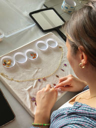 Hands assembling a handmade gold charm necklace on a white cloth at a craft table, enamel charms sorted in small round trays with a handheld mirror and smartphone nearby