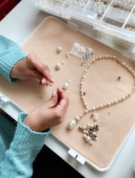 Hands stringing pearls and crystals to assemble a handmade pearl necklace and earring set on a beige jewelry work mat, with compartment box and scattered white and grey beads in a DIY jewelry studio.