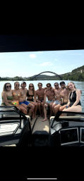 Group of nine friends in swimsuits posing on a motorboat with the Pennybacker (360) Bridge and tree-lined hills over Lady Bird Lake in Austin, Texas on a sunny day — fun summer boating scene.
