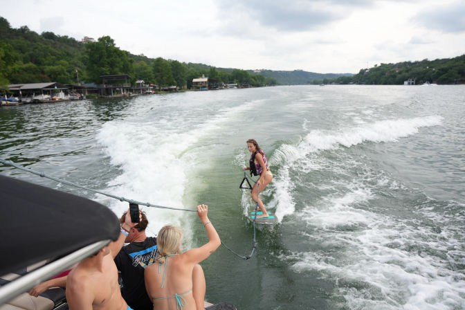 Woman wakesurfing on a lake behind a boat, riding the boat wake past tree-lined shore and boathouses while friends watch from the boat