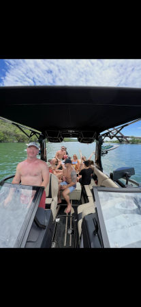 Sun-soaked group of friends on a black-canopied wake boat cruising a green lake near a tree-lined shoreline and bridge, people in swimsuits chatting and relaxing.