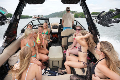 Group of friends in colorful bikinis enjoying a summer boat ride on a lake, laughing aboard a wakeboard boat with a cooler and wakeboard tower, hilly shoreline in the background