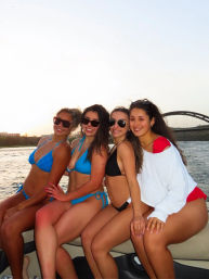 Four friends in colorful bikinis smiling on a boat at sunset, posing together with a river and arched bridge in the background, cheerful summer vibe