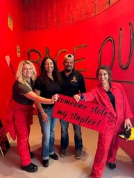 Four smiling adults in a bright red indoor smash (rage) room, two wearing red coveralls holding a red sign that reads "someone stole my stapler!" in front of a curved wall painted with large "RAGE OUT" graffiti.