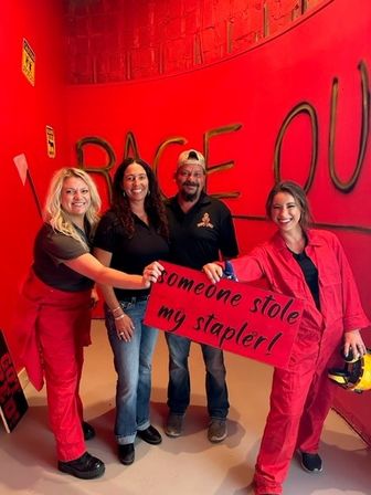 Four smiling adults in a bright red indoor smash (rage) room, two wearing red coveralls holding a red sign that reads "someone stole my stapler!" in front of a curved wall painted with large "RAGE OUT" graffiti.