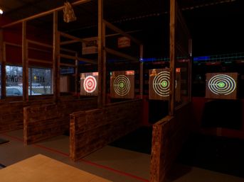 Rustic indoor axe-throwing lanes with wooden partitions and chain-link dividers, glowing electronic bullseye targets projected on wooden backboards in a dim entertainment venue.