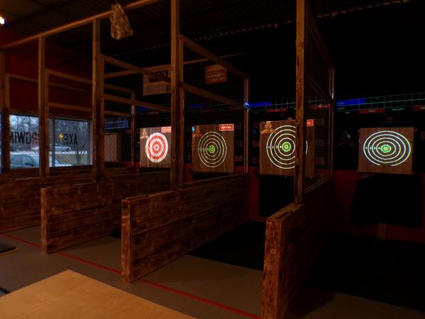 Rustic indoor axe-throwing lanes with wooden partitions and chain-link dividers, glowing electronic bullseye targets projected on wooden backboards in a dim entertainment venue.